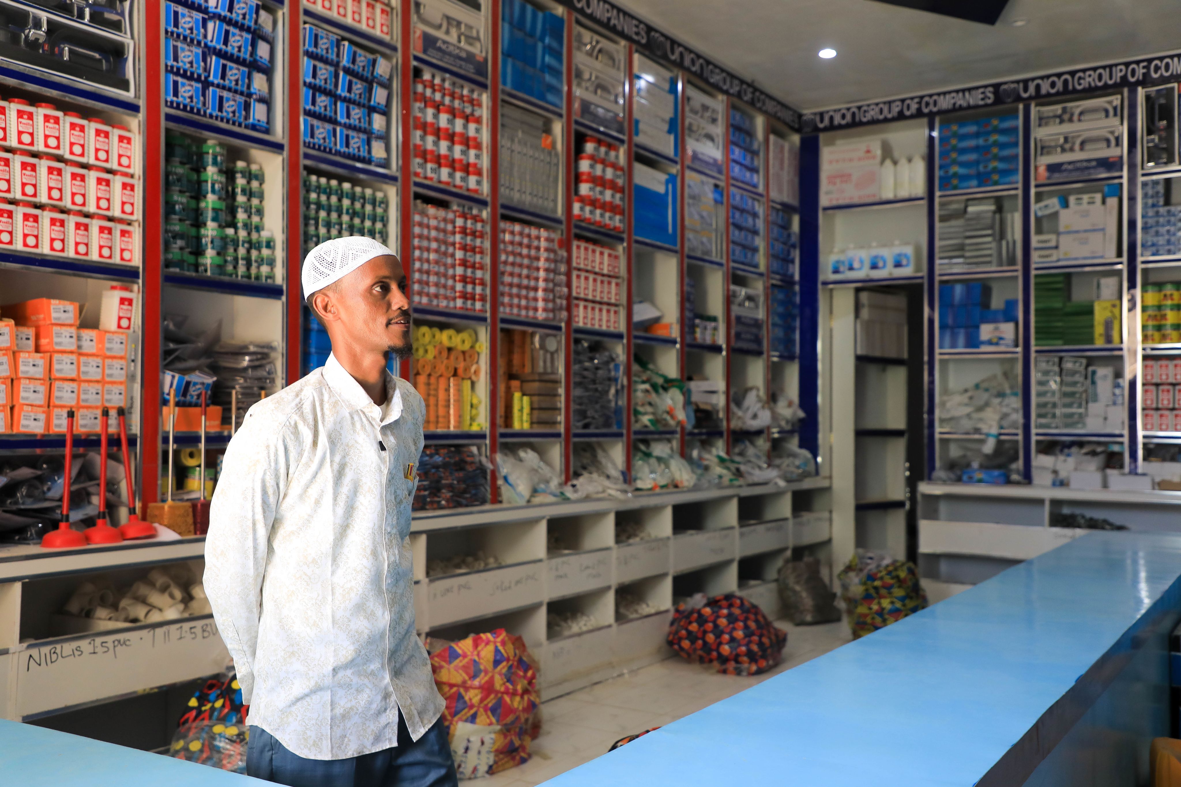 A somali man in a white cap and patterned shirt stands inside a well-stocked hardware store, looking to the side. Shelves filled with neatly arranged boxes of electrical supplies and other hardware items line the walls behind him. The store has a blue and white color scheme and is brightly lit.