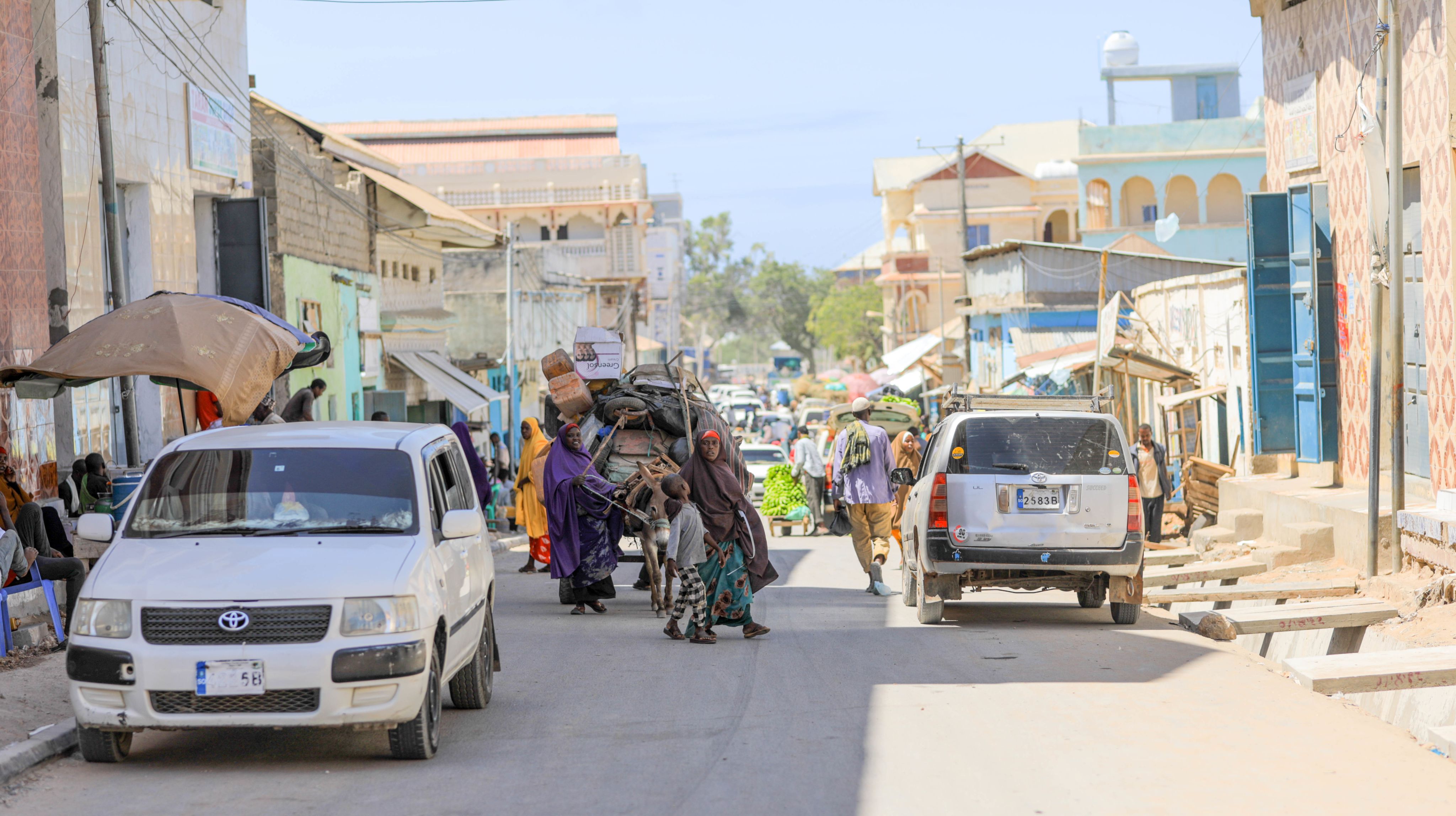 A busy street scene in Somalia with pedestrians and cars on a sunny day, showcasing the activity of a town with shops lining the road.