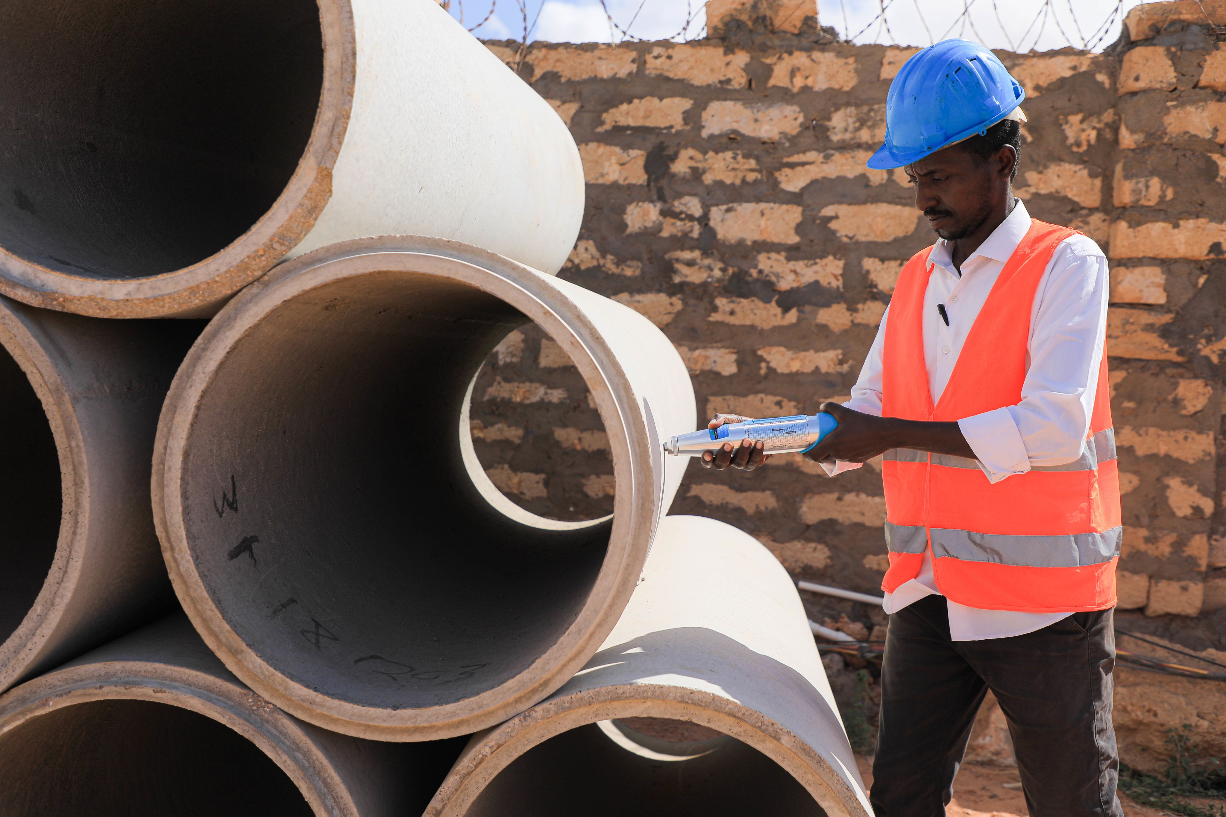  A worker in a blue hard hat and orange high-visibility vest is holding a clipboard, standing beside large concrete pipes with a brick wall in the background.
