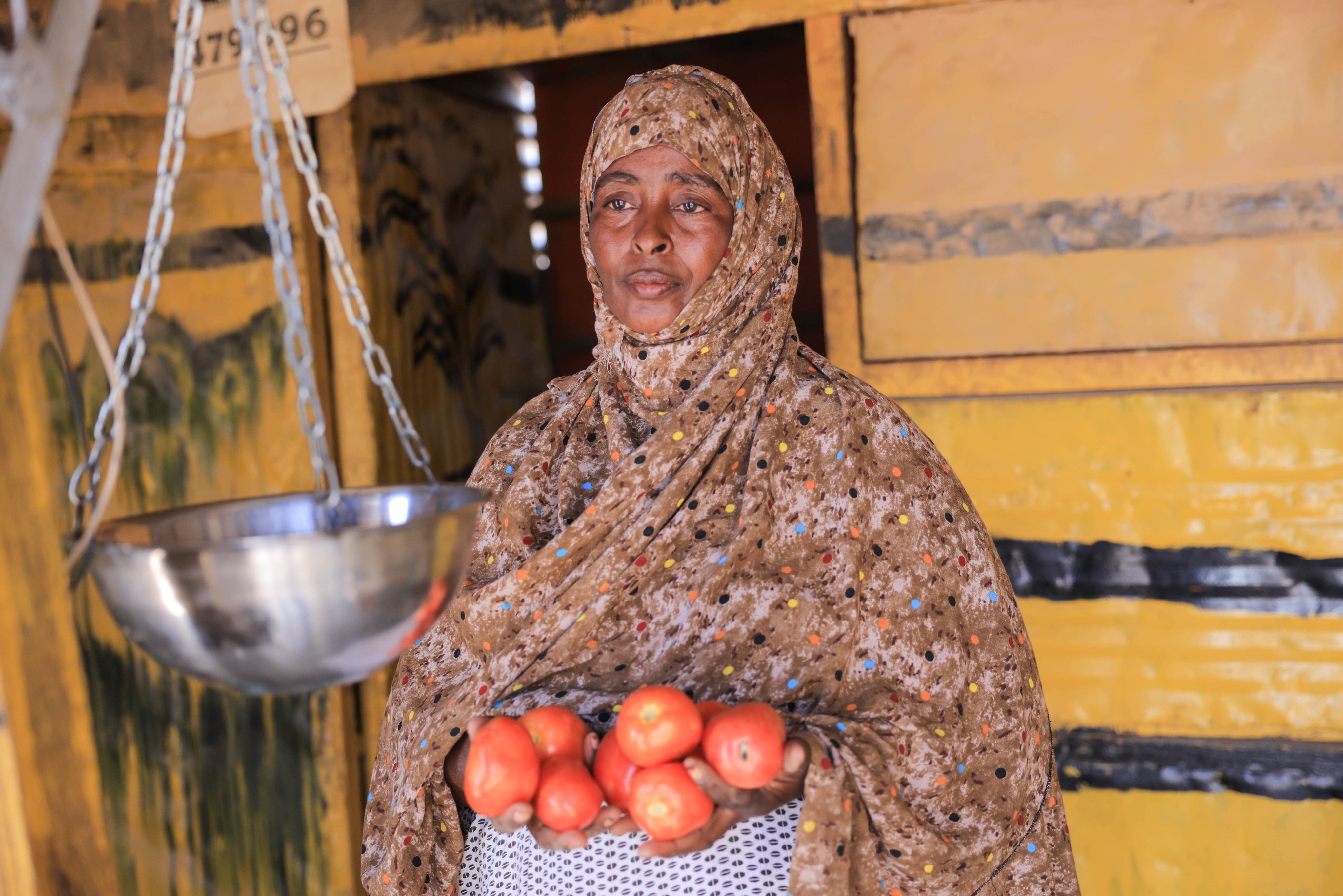  A Somali woman in a patterned headscarf stands in front of a yellow stall with a weighing scale to her side. She is holding a bowl containing several red tomatoes. The background features part of the stall with a rustic, painted surface.