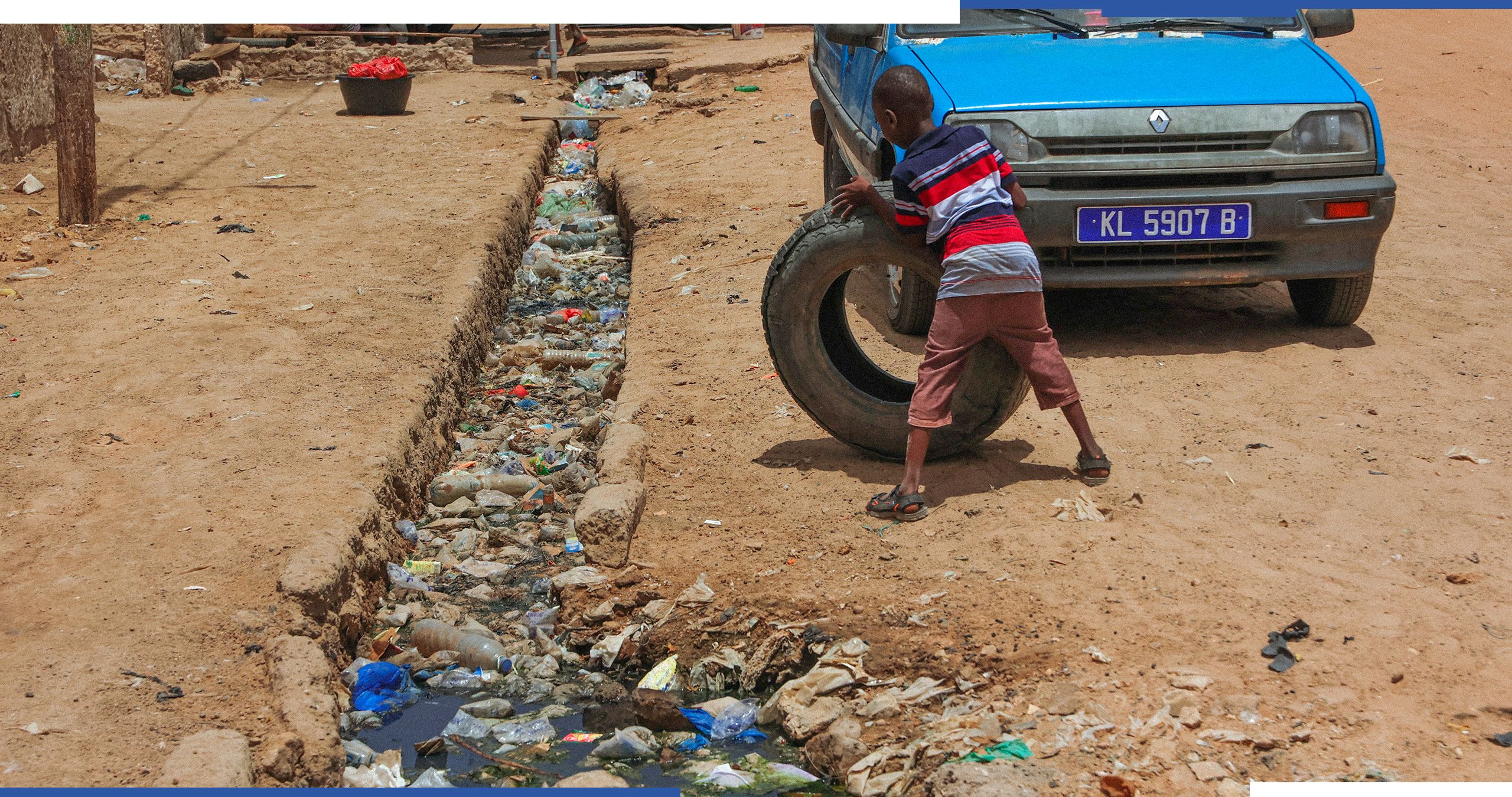 Un niño rodando una rueda junto a un coche y un canal lleno de plásticos flotando en el agua en la ciudad de Kaolack. 